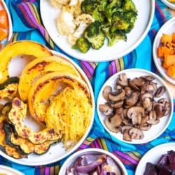 Varying sizes of white round plates on top of a vibrant colorful striped fabric. Each plate is filled with different types of roasted vegetables.