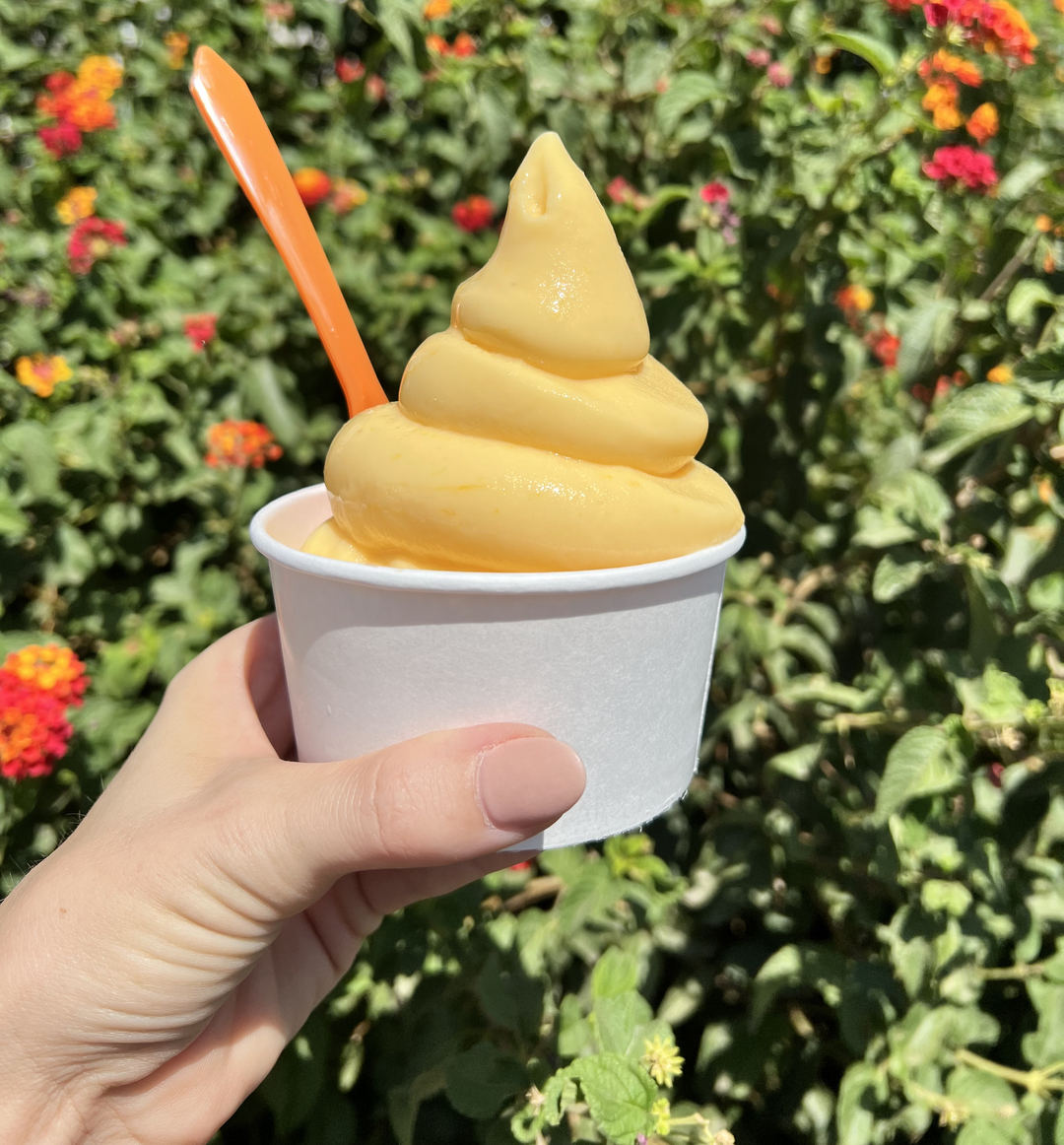 holding a soft serve orange ice cream in front of a flower bush