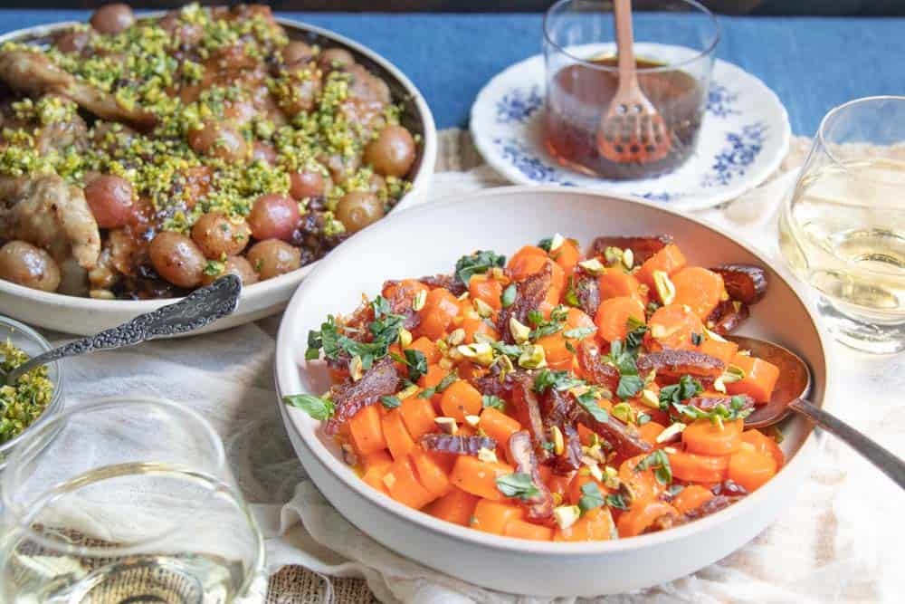 A serving dish of carrots and one of chicken with grapes and raisins, next to a small jar of honey.