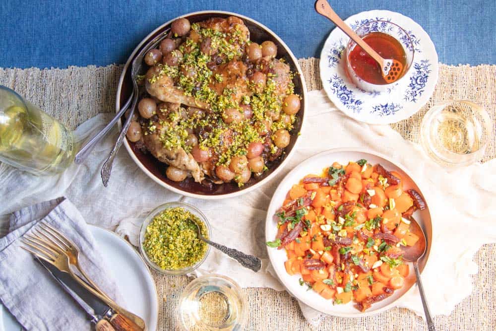 A table set with a dish of Chicken with Wine, Grapes, and Raisins, Hon Glazed Carrots, and glasses of wine.