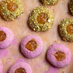Two types of thumbprint cookies with apricot jam on a sheet pan with brown parchment paper. One is covered in pink raspberry sugar and one is rolled in chopped pistachios.