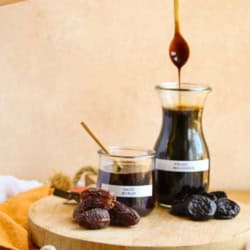 Labeled bottles of date syrup and prune molasses next to their dried fruit counterparts. A wooden spoon is drizzling prune molasses back into the jar after being dipped into it to demonstrate the consistency.