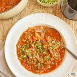 A dinner table with a bowl of cabbage roll soup, a small dish of gremolata for garnish, and red wine in a stemless glass.