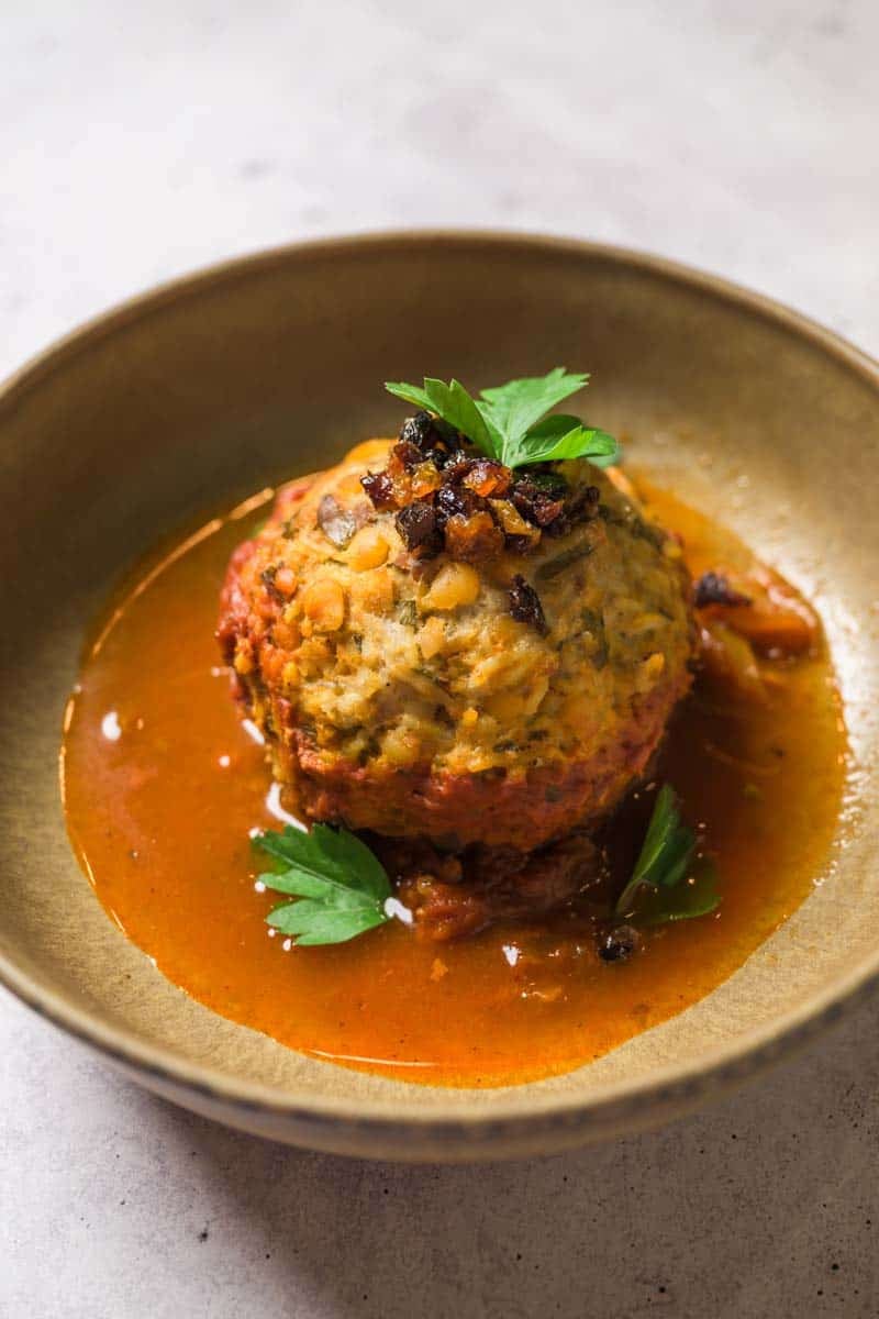 A large meatball sitting in a bowl of red sauce topped with spiced raisins.