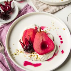 A serving of hibiscus poached pears and apples with unsweetened whipped cream underneath, and a drizzle of pink poaching syrup and green pistachios crumbles.