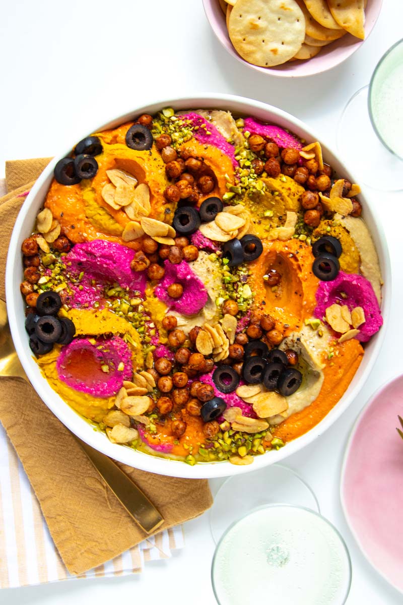 A serving dish filled with dollops of plain, roasted red pepper, and beet hummus layered by spoonfulls onto a serving platter then topped with crispy chickpeas, crispy garlic slices, crushed pistachios, sliced black olives, and seasonings.